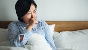 Persona tosiendo, sentada en la cama, llevando pijama de rayas azules. El entorno es un dormitorio con sábanas blancas y cabecera de madera.