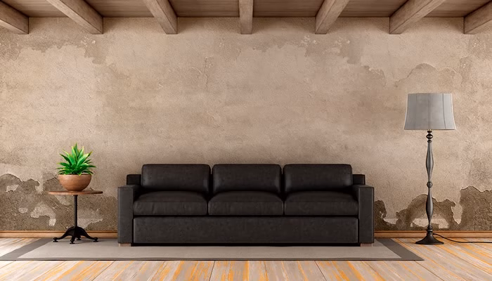 Un sofá negro está situado en el centro. A su izquierda hay una mesa con una planta. A su derecha, una lámpara alta. Fondo de pared desgastada y techo de vigas.