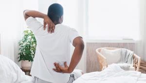 Una persona está sentada en la cama masajeándose la espalda. Alrededor, hay una planta verde y una silla de mimbre junto a una ventana luminosa.