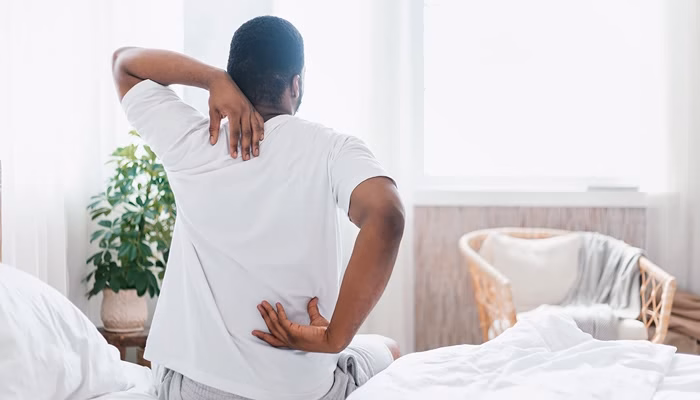 Una persona está sentada en la cama masajeándose la espalda. Alrededor, hay una planta verde y una silla de mimbre junto a una ventana luminosa.