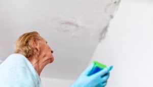 Persona mayor observa el techo con manchas de humedad. Lleva guantes azules y en la mano sostiene una esponja. La pared y el techo son blancos, con áreas oscuras.