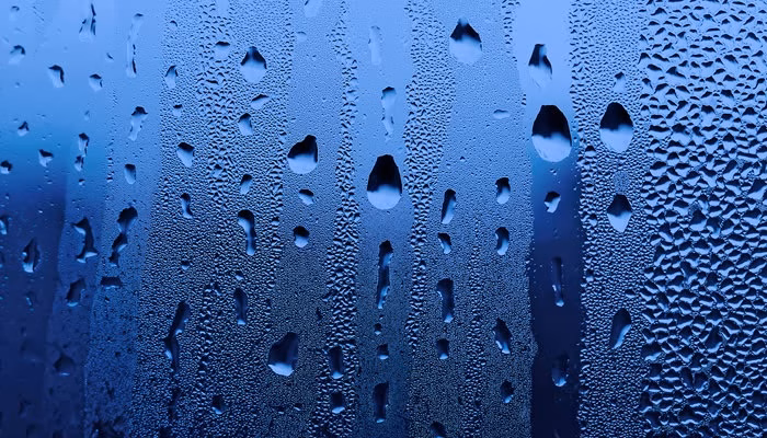 Gotas de agua se deslizan sobre una superficie de vidrio, creando patrones de condensación. El fondo es azul, sugiriendo un día frío o lluvioso en el exterior.
