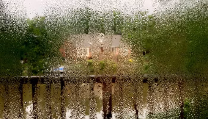 Una ventana cubierta de gotas de agua; llueve intensamente. Fuera, un jardín verde y una casa de ladrillo son visibles. El clima nuboso resalta la humedad.