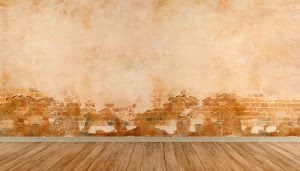 Una pared de ladrillos parcialmente cubierta por yeso desgastado, revelando zonas deterioradas, sobre un suelo de tablones de madera, en un ambiente interior sin muebles visibles.