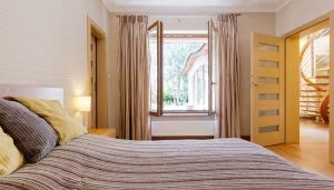 Cama con cojines en tonos marrón y amarillo, ubicada frente a una amplia ventana abierta. El contexto incluye cortinas y puertas que conectan con un pasillo iluminado y escaleras.