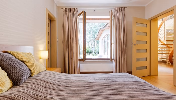 Cama con cojines en tonos marrón y amarillo, ubicada frente a una amplia ventana abierta. El contexto incluye cortinas y puertas que conectan con un pasillo iluminado y escaleras.