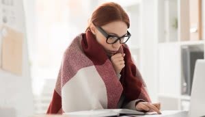 Persona envuelta en una manta, mirando una computadora. Parece concentrada en su trabajo. Está en una oficina iluminada con estanterías y documentos alrededor.