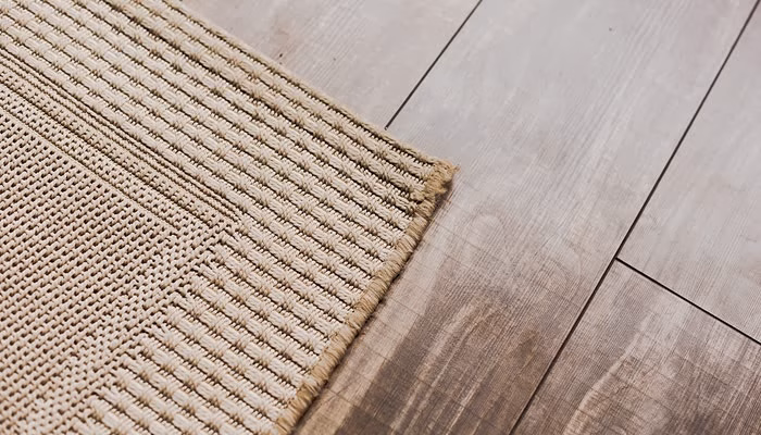Una alfombra de tejido grueso descansa sobre un suelo de madera clara. El diseño presenta líneas entrelazadas en un ambiente luminoso.