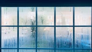 Cristal: ventanas con gotas de condensación. Acción: el agua resbala suavemente. Contexto: enfría la vista exterior de un vecindario residencial con edificios borrosos, visibles a través del vidrio empañado.