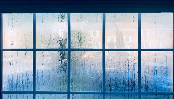Cristal: ventanas con gotas de condensación. Acción: el agua resbala suavemente. Contexto: enfría la vista exterior de un vecindario residencial con edificios borrosos, visibles a través del vidrio empañado.