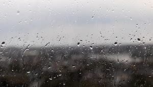 Gotas de lluvia resbalan por un cristal. Tras el vidrio, un paisaje urbano se ve borroso bajo un cielo nublado. No hay texto visible.