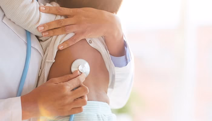 Un médico escucha el pecho de un niño con un estetoscopio. El niño está en brazos, mostrando un ambiente de atención médica cálida y luminosa.