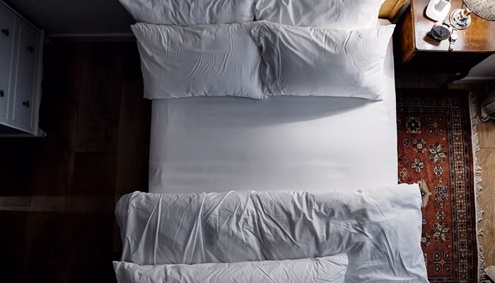 Una cama blanca perfectamente hecha con almohadas alineadas, ubicada en un dormitorio. A su lado, hay una mesita de noche con objetos y una alfombra decorativa en el suelo.