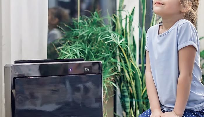 Un purificador de aire en funcionamiento muestra "54" en su pantalla, mientras una niña sentada cerca observa. Al fondo, hay plantas y una ventana.