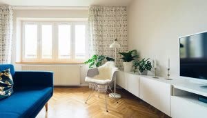 Un sofá azul se encuentra en una sala de estar luminosa, junto a una silla con manta, plantas, lámpara y un televisor sobre un mueble blanco. Ventanales iluminan el espacio.