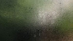 Cristales empañados, con gotas de agua deslizándose y reflejando luz, crean un efecto borroso. El fondo muestra un paisaje verde, sugiriendo un ambiente exterior húmedo y nublado.