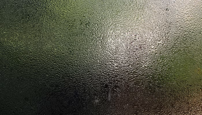Cristales empañados, con gotas de agua deslizándose y reflejando luz, crean un efecto borroso. El fondo muestra un paisaje verde, sugiriendo un ambiente exterior húmedo y nublado.