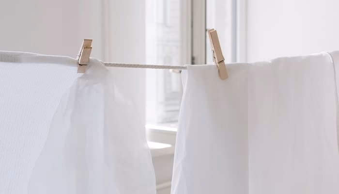 Dos pinzas de madera cuelgan ropa blanca en una cuerda, iluminada por luz natural, cerca de una ventana en un interior.