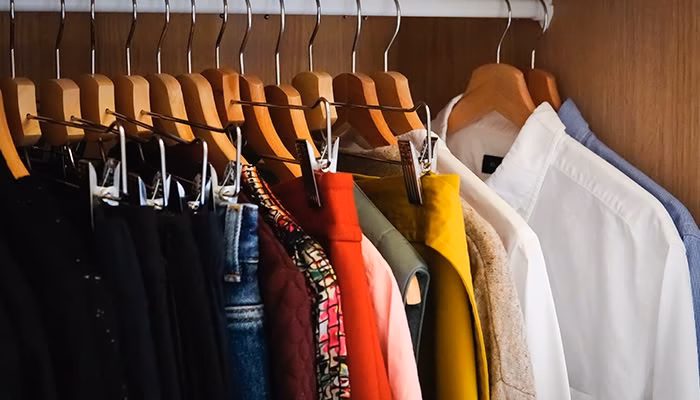 Perchas de madera sostienen ropa variada, incluyendo camisetas y chaquetas de diferentes colores. Están colgadas ordenadamente dentro de un armario de madera. No hay texto visible.