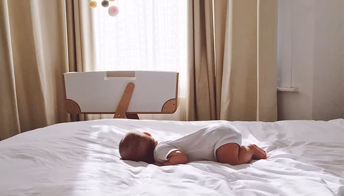 Un bebé reposa boca abajo sobre una cama blanca. Al fondo, hay una cuna junto a una ventana cubierta con cortinas claras, creando un ambiente tranquilo.