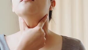Una persona toca su garganta con una mano, posiblemente indicando molestias o dolor. El contexto es un interior iluminado de manera suave, con fondo desenfocado.