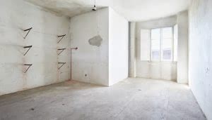 Habitación vacía con paredes y suelo de cemento. Tubería de agua expuesta y repisas metálicas en la pared izquierda. Luz entrando por una gran ventana con contraventanas abiertas.