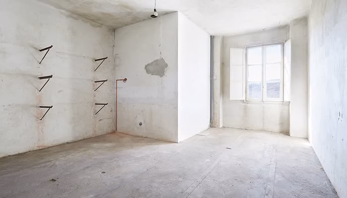 Habitación vacía con paredes y suelo de cemento. Tubería de agua expuesta y repisas metálicas en la pared izquierda. Luz entrando por una gran ventana con contraventanas abiertas.