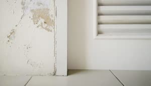 Una pared dañada está al lado de una puerta blanca con persianas. El suelo es de baldosas blancas, creando un ambiente interior simple y descuidado.