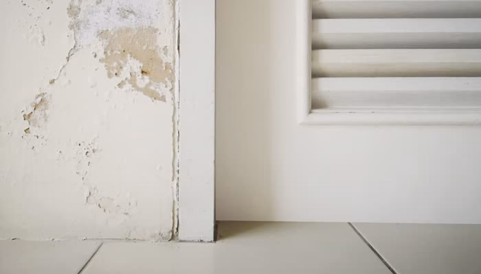 Una pared dañada está al lado de una puerta blanca con persianas. El suelo es de baldosas blancas, creando un ambiente interior simple y descuidado.