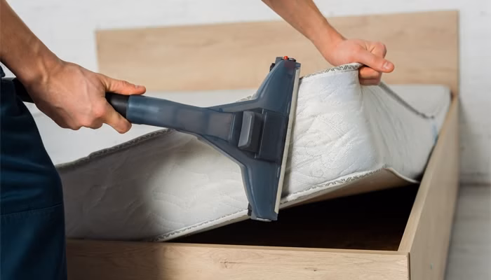 Una mano sostiene una aspiradora mientras limpia un colchón levantado en un marco de cama de madera, probablemente en un dormitorio, sugiriendo tareas de limpieza del hogar.