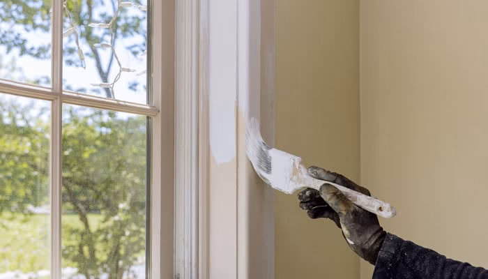 Un pincel pinta un marco de ventana de blanco. Una mano con guante sostiene el pincel. Afuera, se ve un árbol verde a través del vidrio.