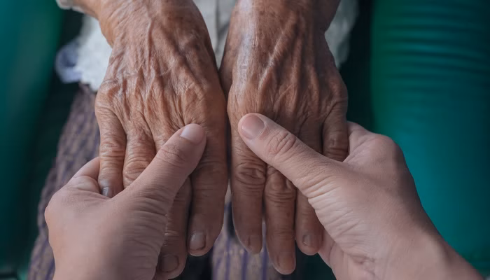 Manos envejecidas, son sujetadas suavemente por manos más jóvenes, en un gesto de apoyo y cuidado, sobre un fondo de asiento verde.