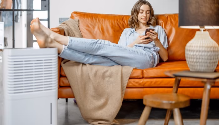 Una persona recostada en un sofá naranja, usando un teléfono. Lleva ropa cómoda. A su lado hay un deshumidificador y una mesa con una lámpara, creando un ambiente acogedor.