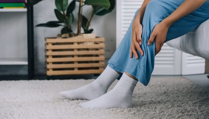 Una persona sentada en una cama sostiene su pierna, vistiendo pantalón azul y calcetines blancos, en una habitación iluminada con alfombra clara y planta en maceta.