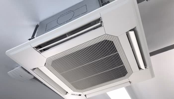 Unidad de aire acondicionado de cassette instalada en el techo, emitiendo aire fresco, en una habitación de paredes y techo blanco con iluminación fluorescente.