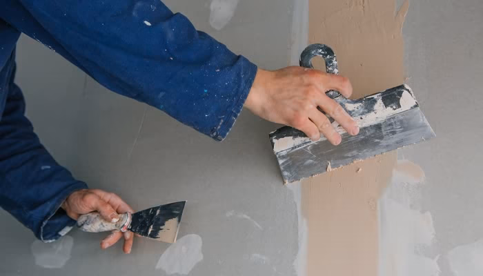 Manos usando espátulas aplican yeso sobre una pared. La acción está en un contexto de trabajo de construcción o reparación, con herramientas cubiertas de material blanco.
