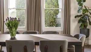 Un ramo de flores moradas adorna una mesa de comedor rectangular en una sala iluminada por dos ventanas amplias con cortinas beige. Una planta alta está en una esquina.