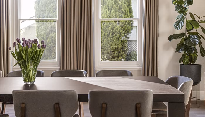 Un ramo de flores moradas adorna una mesa de comedor rectangular en una sala iluminada por dos ventanas amplias con cortinas beige. Una planta alta está en una esquina.