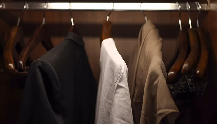 Ropa colgada en perchas de madera, dentro de un armario. Una chaqueta negra y dos camisetas, una blanca y una beige, organizadas ordenadamente. Fondo marrón del armario.