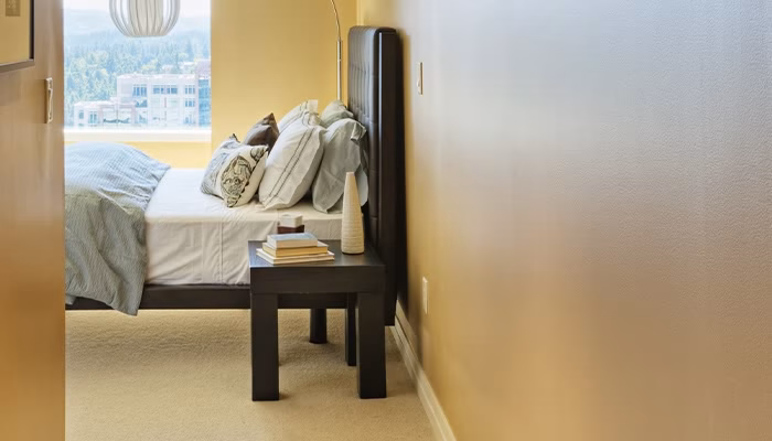 Cama con sábanas blancas y cojines decorativos, situada al lado de una ventana luminosa. Mesa de noche con libros y decoración en una habitación de paredes beige.