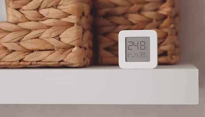 Un termómetro digital muestra "24.8°C" y "(^) 39%" en una estantería blanca, junto a cestas de mimbre entrelazadas.