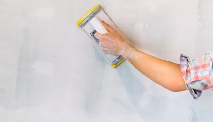 Una mano sostiene una lijadora manual, que suaviza una pared blanca. La persona lleva una camisa de cuadros, y parece estar renovando o reparando la superficie.