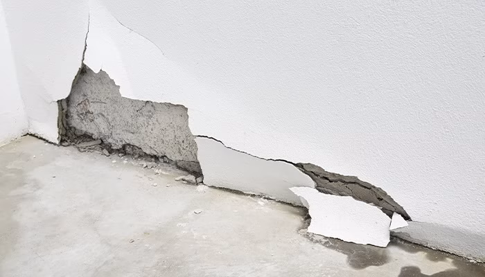 Una pared presenta un gran agrietamiento en la superficie, exponiendo el material interno. Está situada sobre un suelo de concreto en un entorno interior.