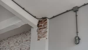 Un muro interior con ladrillos expuestos en una esquina, rodeado de paredes pintadas de blanco. Tubos eléctricos recorren el techo, y una luz de seguridad está instalada en la pared.