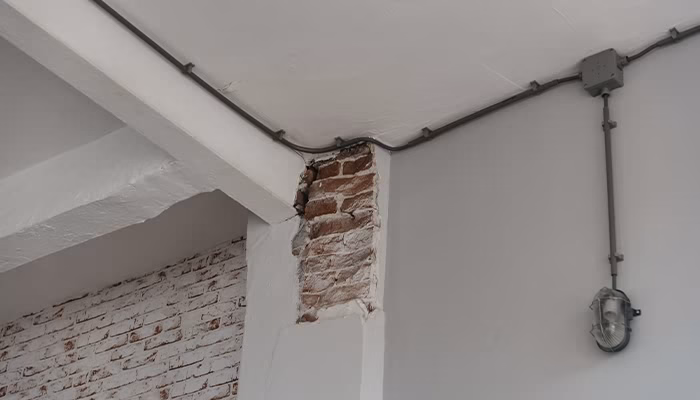 Un muro interior con ladrillos expuestos en una esquina, rodeado de paredes pintadas de blanco. Tubos eléctricos recorren el techo, y una luz de seguridad está instalada en la pared.