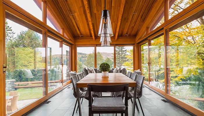 Mesa de comedor rodeada de sillas grises, bajo una lámpara colgante, en una habitación con paredes de vidrio que muestra un paisaje natural exterior con árboles y montañas.