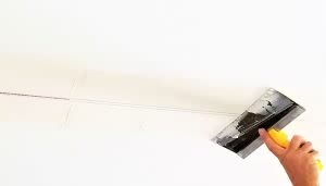 Una mano sostiene una espátula aplicando masilla en una unión de paneles de yeso en el techo, en un entorno interior blanco y bien iluminado.