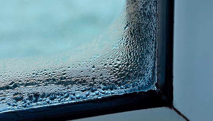 Condensación cubre un vidrio, acumulándose en gotas pequeñas. El marco es oscuro y angulado, creando un contrapunto entre la superficie húmeda y el frío ambiente exterior.