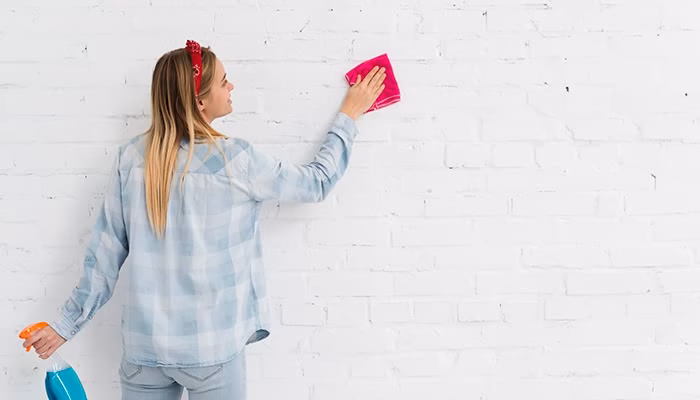 Una persona limpia una pared blanca de ladrillos con un paño rosa, mientras sostiene un rociador azul en una habitación luminosa.