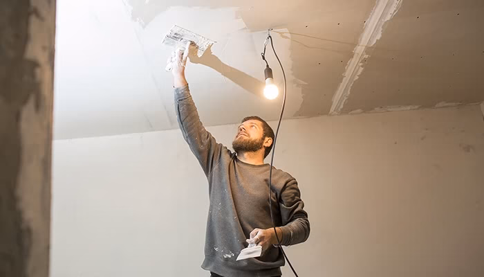 Un hombre repara el techo con yeso, sosteniendo una espátula. A su alrededor, una bombilla cuelga iluminando la habitación en proceso de renovación.